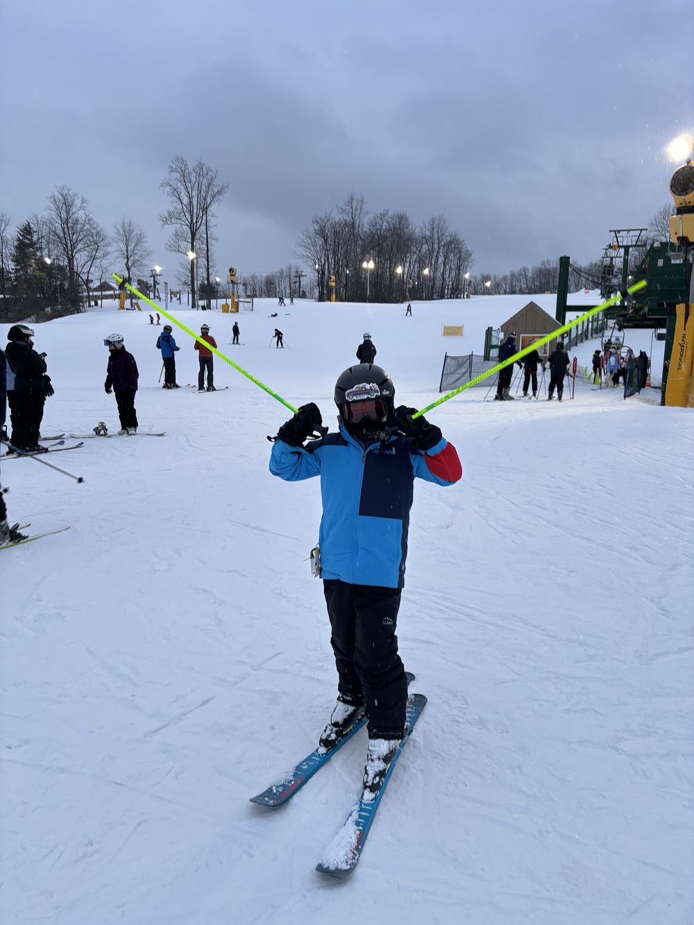 A student at the base area holding ski poles across their shoulders, lift towers behind.